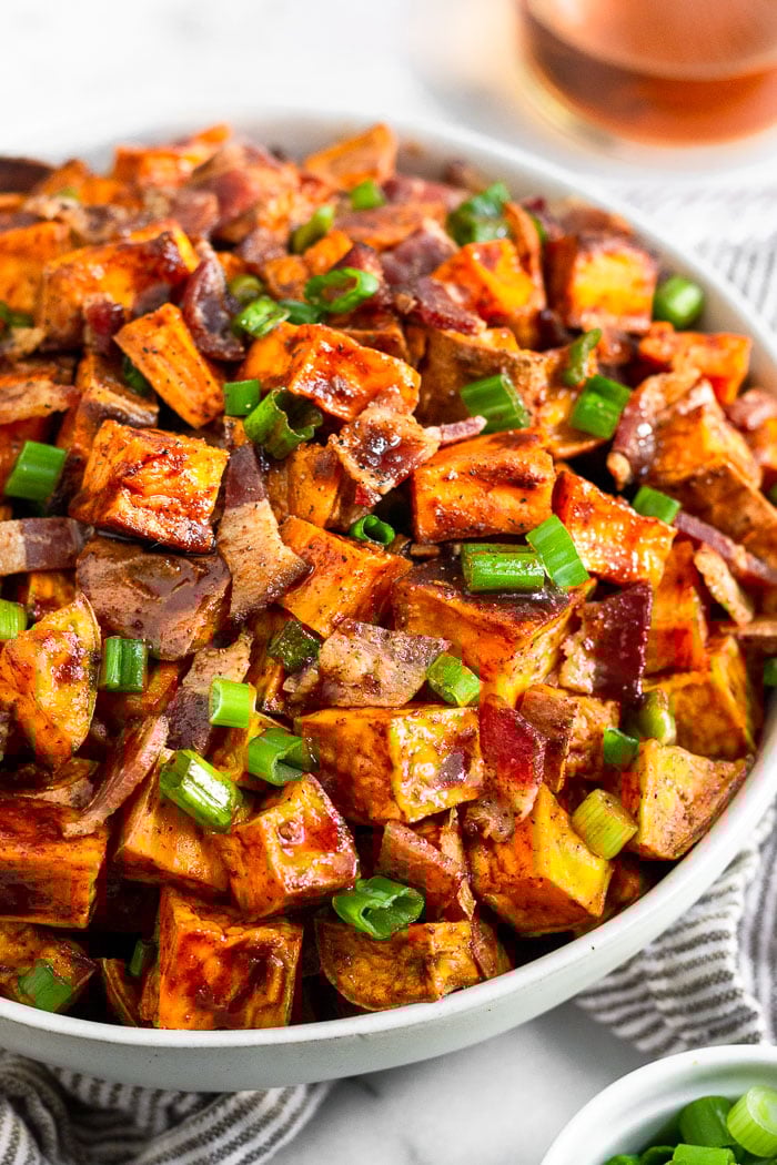 Bowl of glazed sweet potatoes topped with bacon and green onions. Next to is a bowl of green onions and behind it a glass of whiskey.