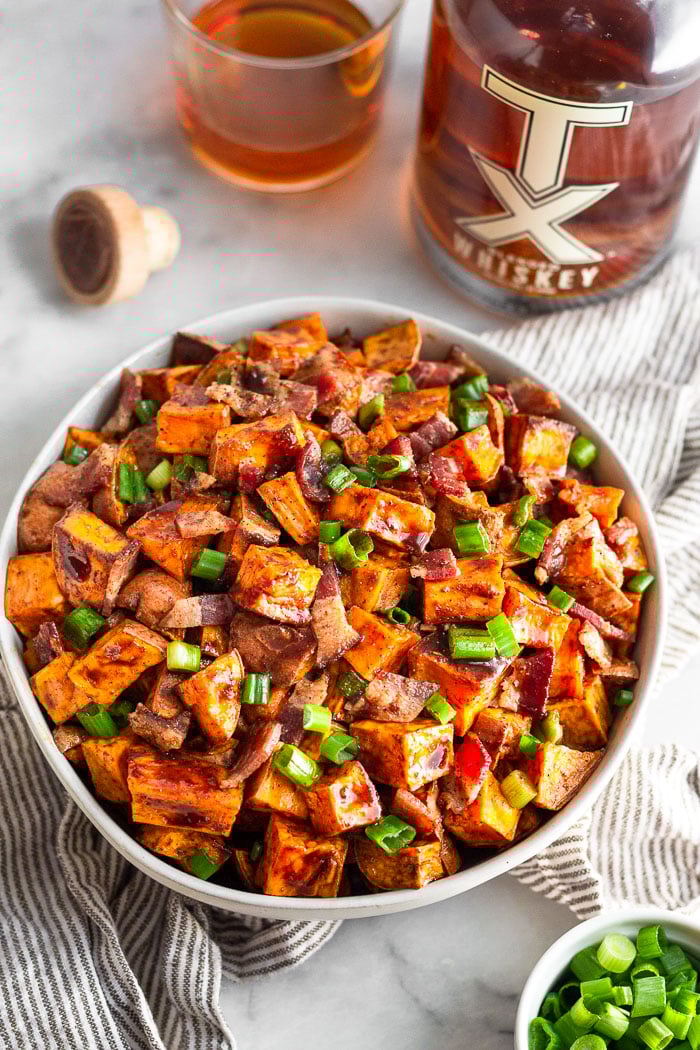 Bowl of whiskey glazed sweet potatoes topped with bacon and green onion. The bowl is sitting on a striped towel with a bowl of green onions, bottle of whiskey, a cup of whiskey, and the bottle's cork next to it. 