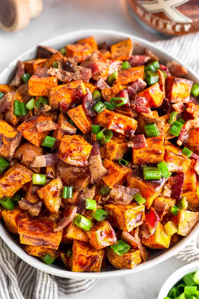 Bowl of whiskey glazed sweet potatoes topped with bacon and chopped green onions. It is sitting on a striped towel with a bowl of green onions and a bottle whiskey around it.