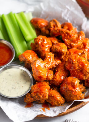 A plate of crispy air fryer buffalo cauliflower covered in buffalo sauce. Also on the plate is cut up celery, a ramekin of ranch, and a ramekin of buffalo sauce.