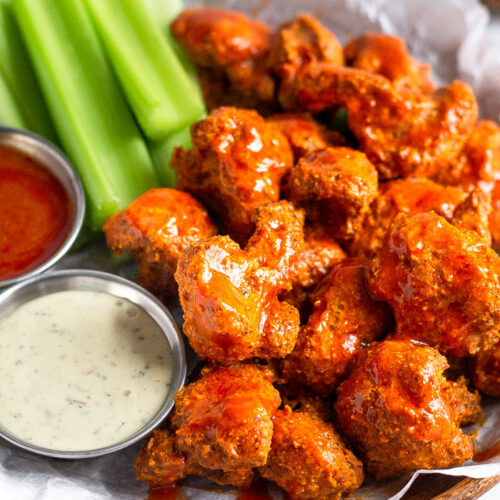 A plate of crispy air fryer buffalo cauliflower covered in buffalo sauce. Also on the plate is cut up celery, a ramekin of ranch, and a ramekin of buffalo sauce.