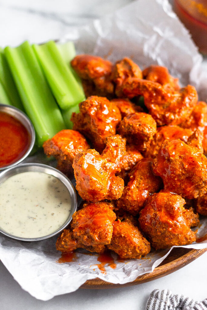 A plate of crispy air fryer buffalo cauliflower covered in buffalo sauce. Also on the plate is cut up celery, a ramekin of ranch, and a ramekin of buffalo sauce.