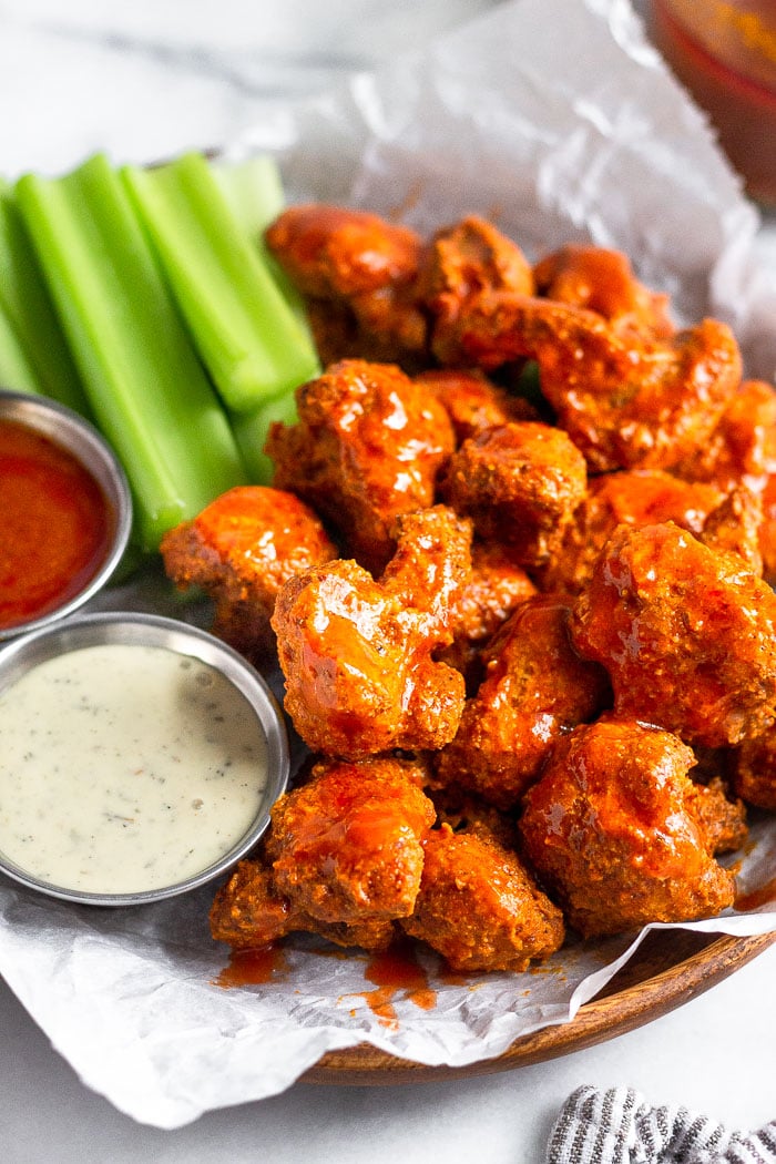 A plate of crispy air fryer buffalo cauliflower covered in buffalo sauce. Also on the plate is cut up celery, a ramekin of ranch, and a ramekin of buffalo sauce.