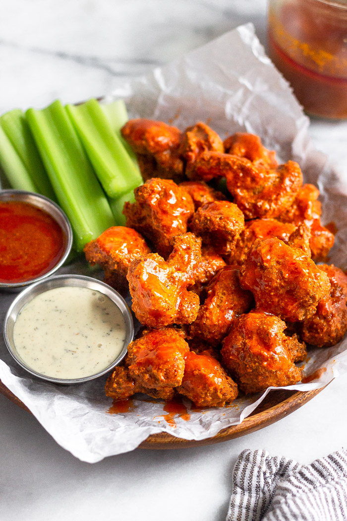 Wooden plate with parchment paper on it with buffalo cauliflower made in the air fryer on it. There is also celery sticks and small dishes of ranch and hot sauce.