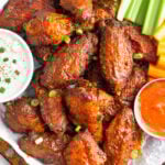 Overhead shot of a pile of air fryer buffalo wings on a baking sheet garnished with chopped green onions. Next to them in are two bowls of blue cheese dressing and hot sauce with celery and carrots on the side.