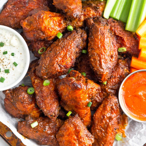Overhead shot of a pile of air fryer buffalo wings on a baking sheet garnished with chopped green onions. Next to them in are two bowls of blue cheese dressing and hot sauce with celery and carrots on the side.