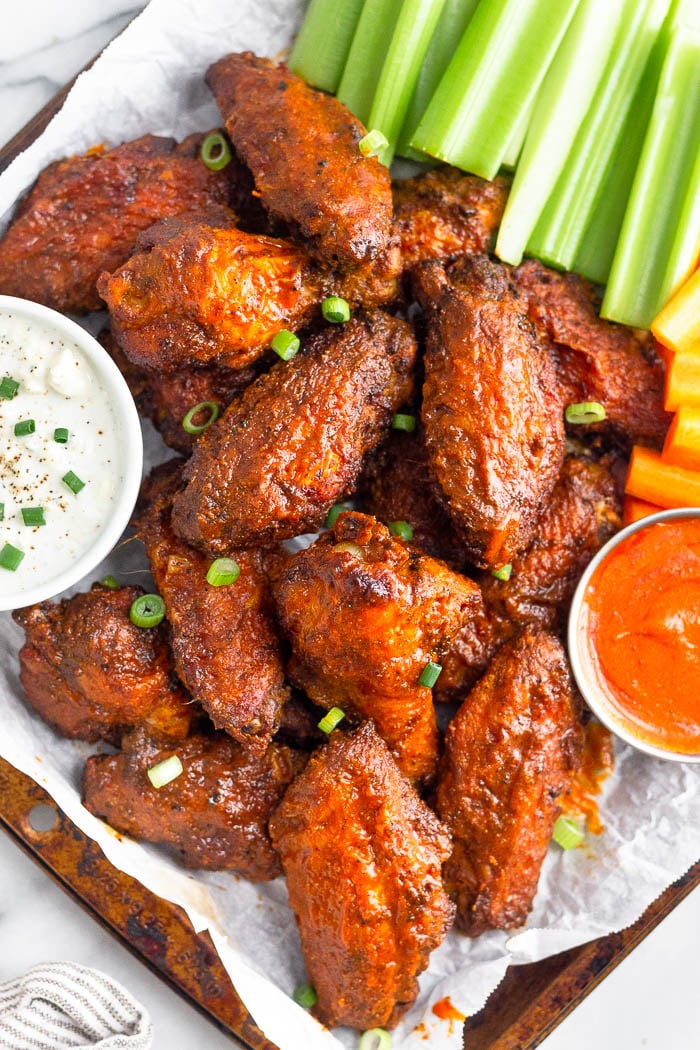 Overhead shot of a pile of air fryer buffalo wings on a baking sheet garnished with chopped green onions. Next to them in are two bowls of blue cheese dressing and hot sauce with celery and carrots on the side.