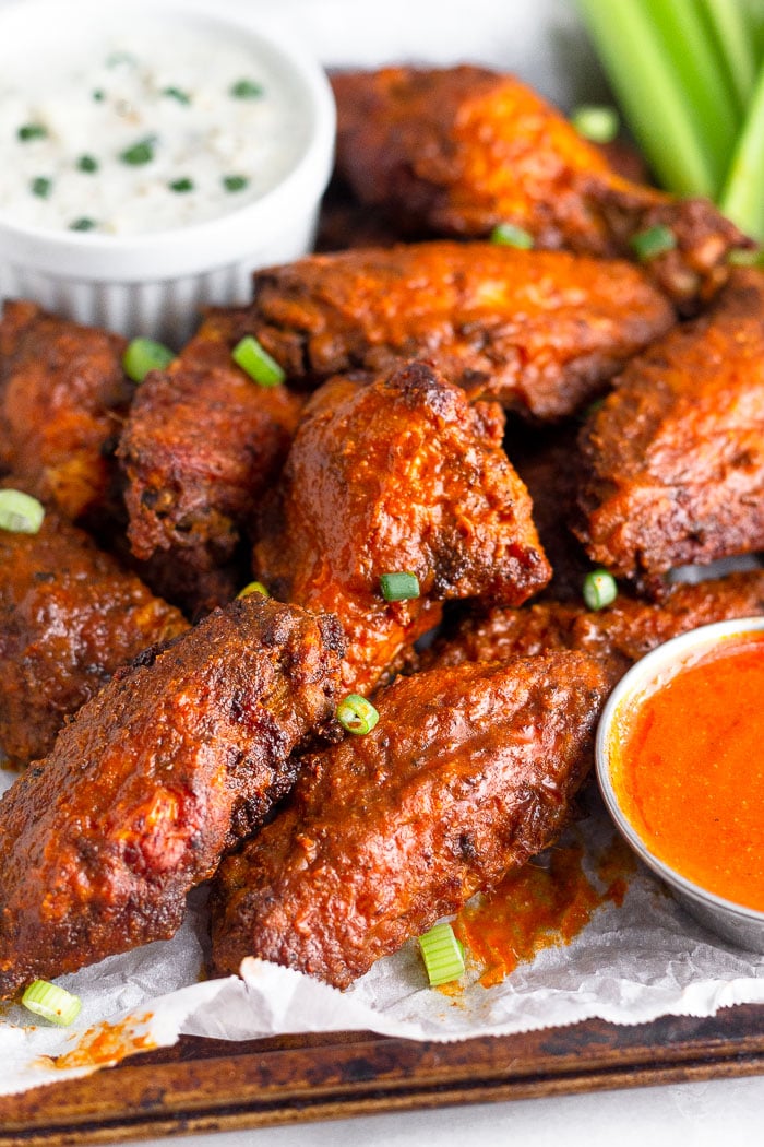 Baking sheet lined with parchment paper with a pile of buffalo chicken wings. There is a small dish of hot sauce towards the front of the pile and a small bowl of blue cheese dressing behind the pile with celery in the corner.