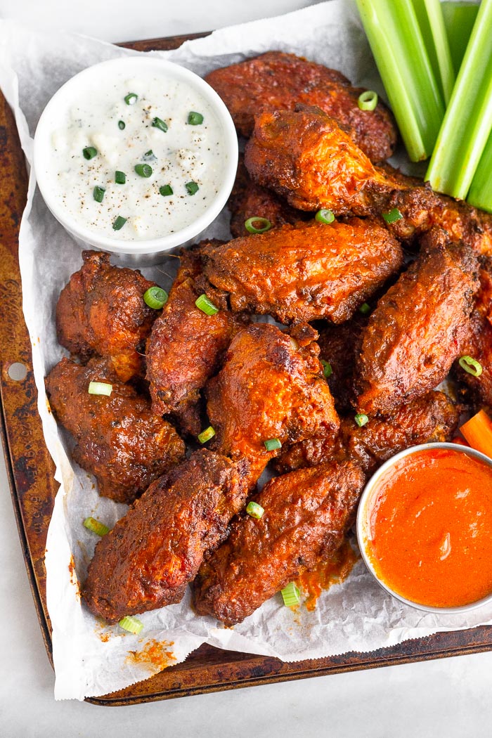 A bunch of air fryer buffalo wings stacked on top of each other on a baking sheet. They are garnished with green onions and next to them is blue cheese and hot sauce for dipping. On the side is celery.