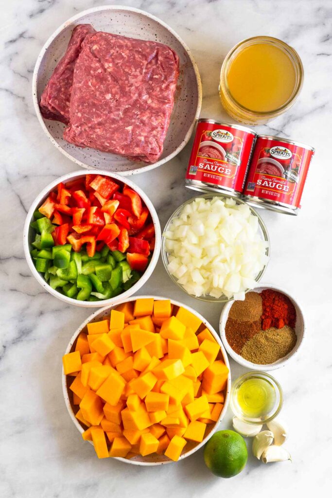White counter with a plate with raw ground meat, a jar of broth, two cans of tomato sauce, a bowl of diced onions, a bowl of spices, a bowl of oil, a few cloves of garlic, a whole lime, a bowl of diced butternut squash, and a bowl of diced bell peppers.