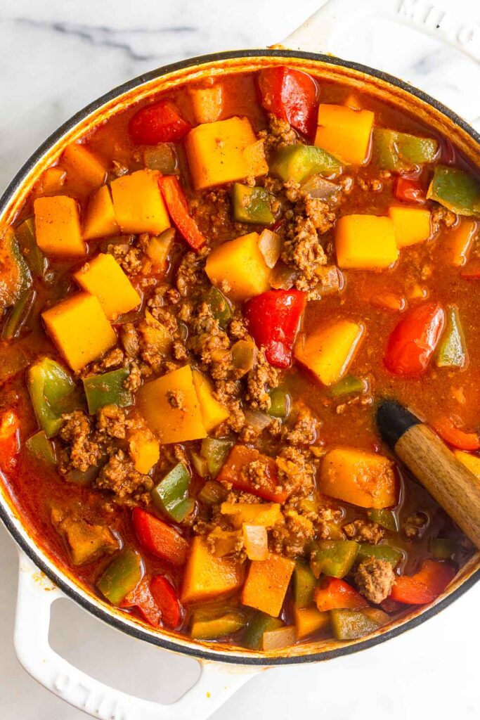Large dutch oven filled with chili with ground buffalo, butternut squash, and veggies. There is a spoon sticking out of the pot.