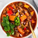 Bison chili in a bowl topped with cilantro and jalapeños with a spoon in it. Another it are some lime wedges, another bowl of chili, and dish of cilantro.