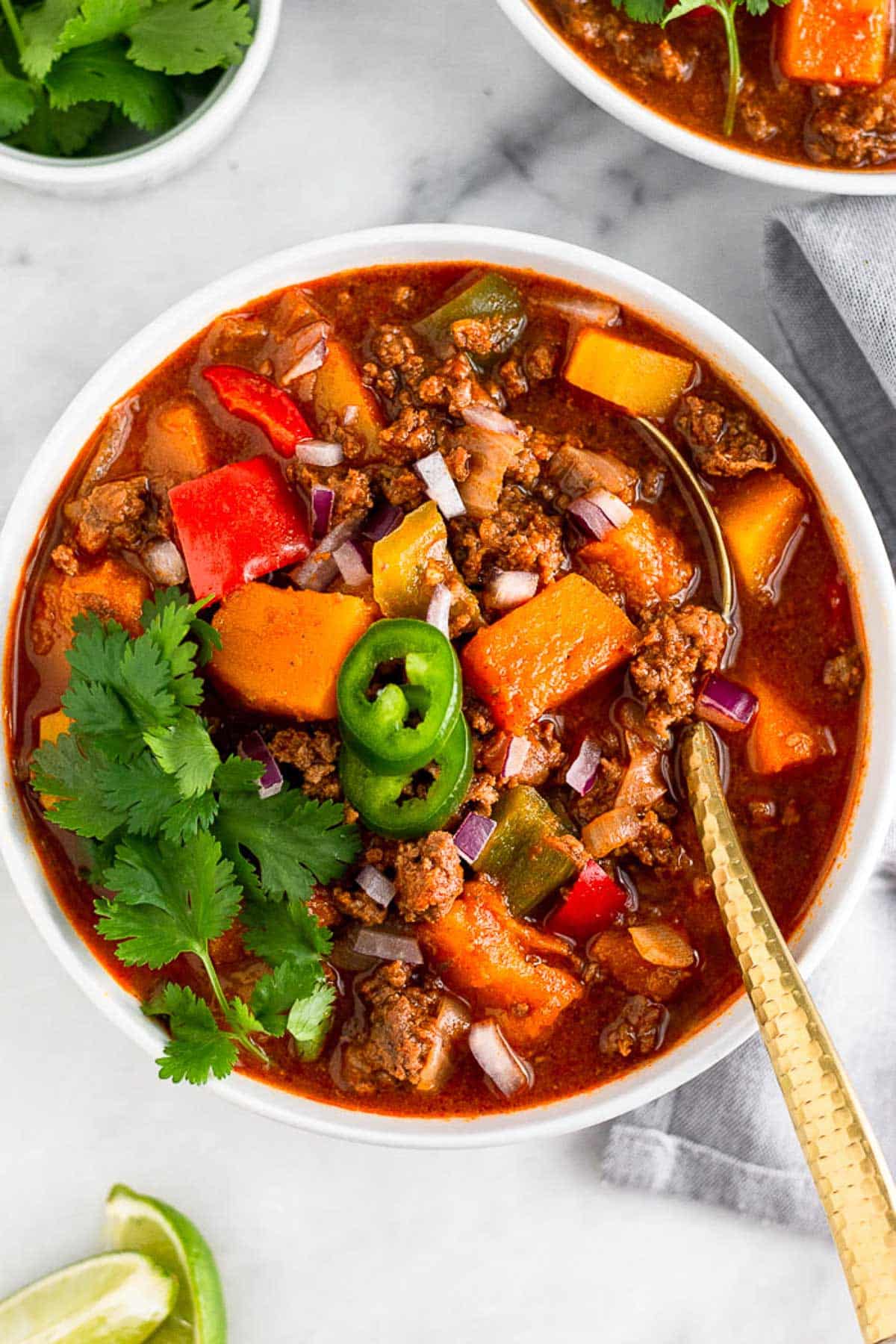 Bison chili in a bowl topped with cilantro and jalapeños with a spoon in it. Another it are some lime wedges, another bowl of chili, and dish of cilantro.
