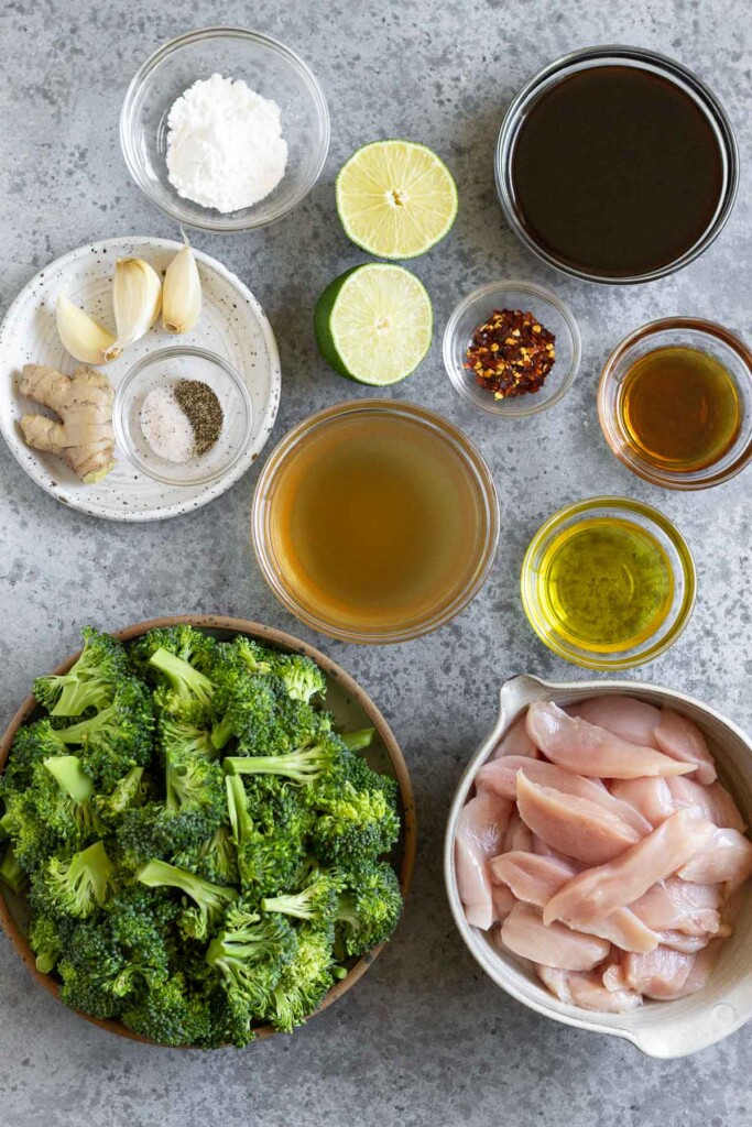 Grey counter with a bowl of coconut aminos, a bowl of red pepper flakes, a bowl of sesame oil, a bowl of olive oil, a bowl of thinly sliced bite-sized pieces of chicken, a bowl of broccoli florets, a bowl of chicken broth, a small plate with a knob of ginger, 3 cloves of garlic, and a bowl of salt and pepper, a lime cut in half, and a small bowl of flour.