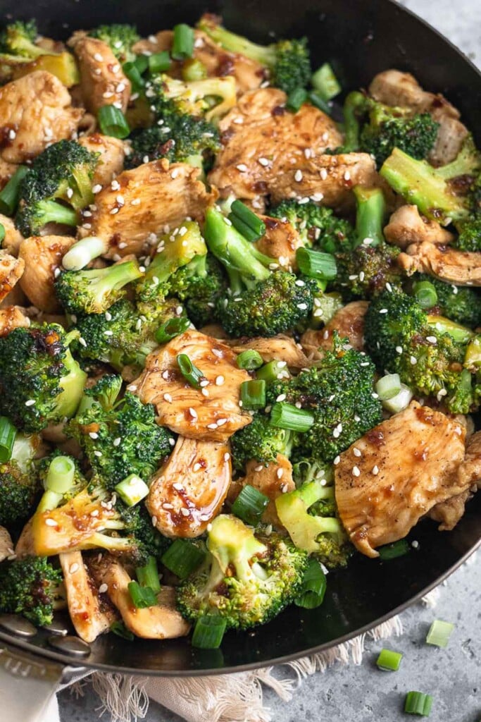 Close up of a pan filled with Chinese chicken and broccoli topped with sliced green onions and sesame seeds.