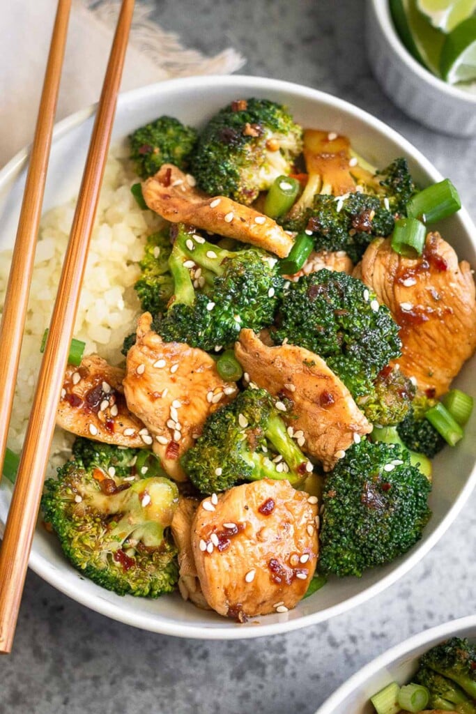 Healthy chicken and broccoli stir fry with white rice in a white bowl. It is topped with sliced green onions and sesame seeds and a a pair of wooden chop sticks is sitting on the bowl. Around the bowl is another bowl of stir fry, a tan linen, and a bowl of lime wedges.
