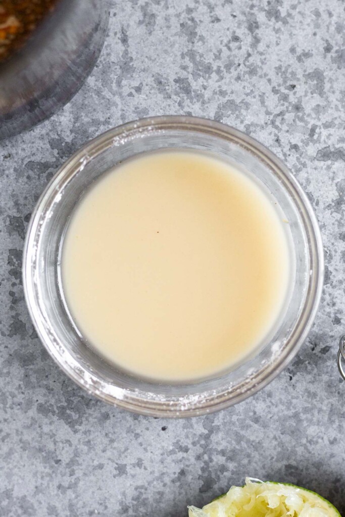 Small bowl filled with a slurry of water and tapioca flour. Next to it is half a juiced lime and a jar filled with stir fry sauce.