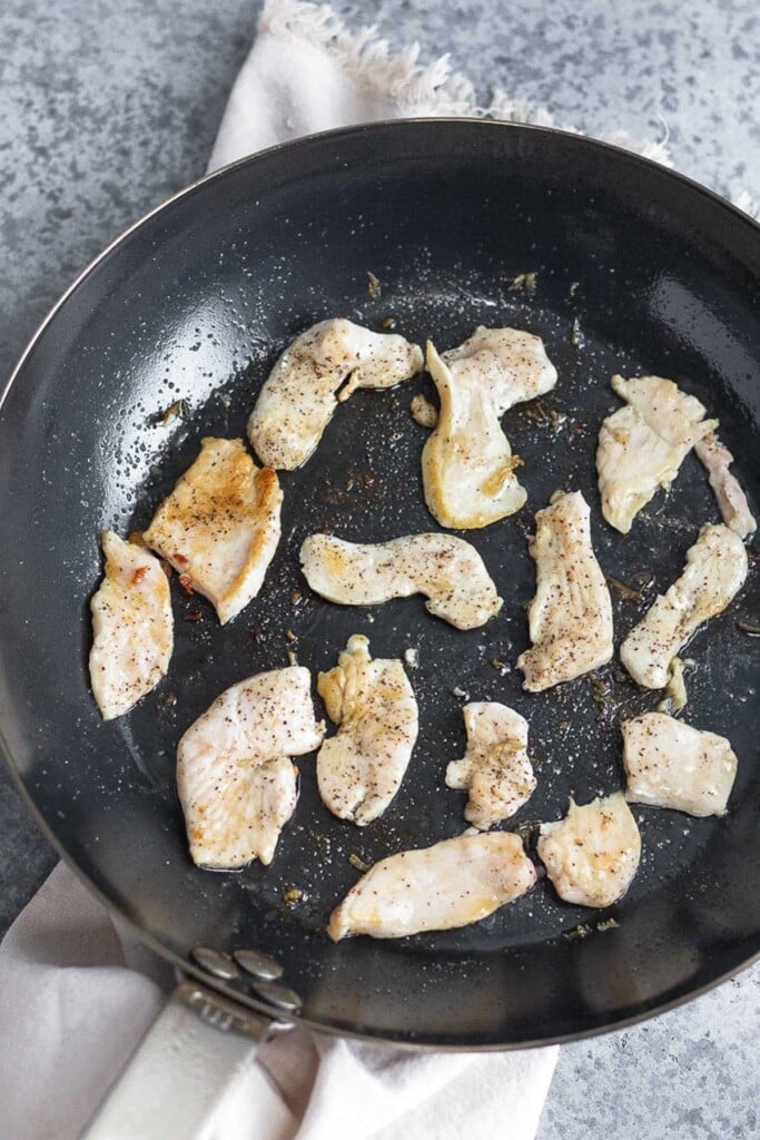 Large skillet filled with thinly sliced bite sized pieces of sautéed chicken. The pan is sitting on a tan linen.