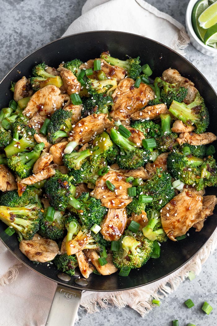 Large skillet filled with Chinese chicken and broccoli stir fry topped with sesame seeds. The pan is on a tan linen and there is a bowl of lime wedges in the corner.