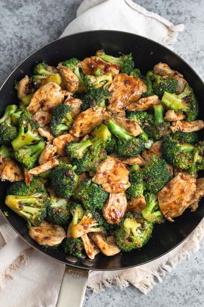 Large pan of sautéed sliced chicken and broccoli tossed with a stir fry sauce. The pan is sitting on a tan linen.