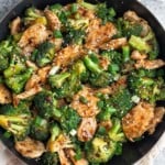Overhead shot of a large skillet filled with chicken and broccoli stir fry. It is topped with sliced green onions and sesame seeds. The pan is sitting on a tan linen and next to it a bowl of lime wedges.