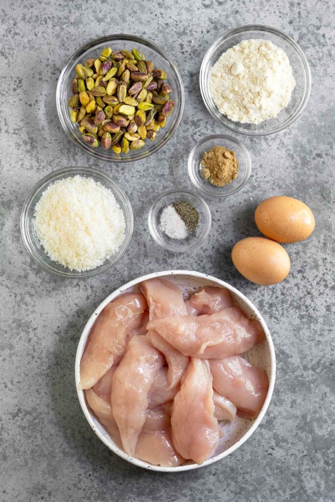 Grey countertop with a bowl of coconut flour, a bowl of ground ginger, a bowl of salt and pepper, two eggs, a plate of raw chicken tenderloins, a bowl of shredded coconut, and a bowl of shelled pistachios.