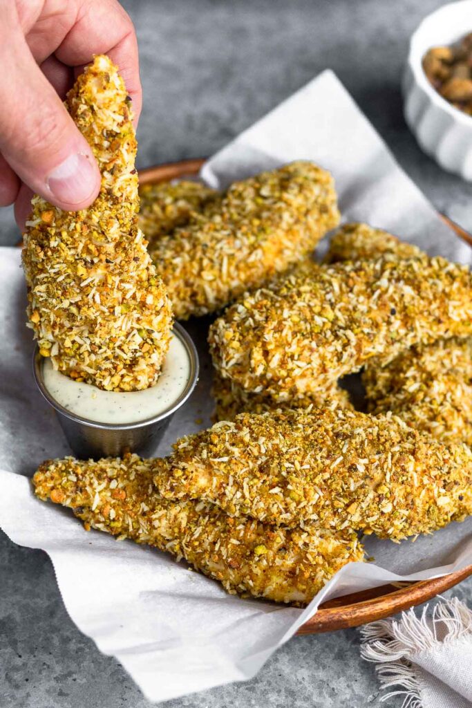 A plate of pistachio crusted chicken tenders with a small dish of ranch. One of the chicken tenders is being dipped in the ranch. Off in the background is bowl of shelled pistachios.