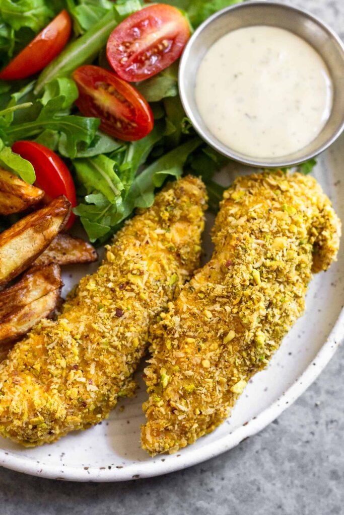 Close up of a plate of baked pistachio chicken tenders with a salad, side of dressing, and french fries.