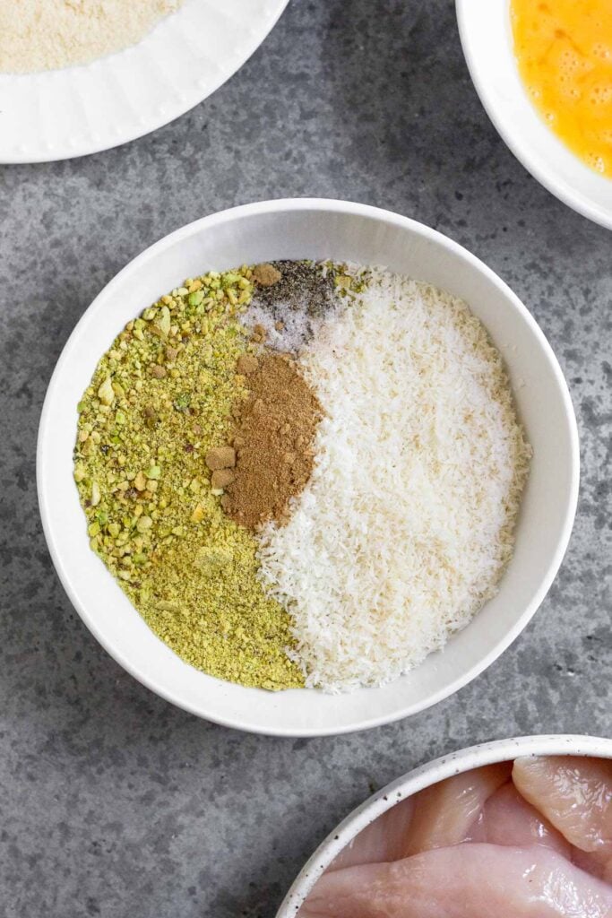 A bowl of ground up pistachios, shredded coconut, and spices. Around it is a plate of raw chicken, a plate with flour on it, and a bowl with whisked eggs.