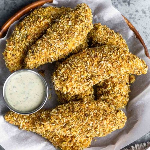 Pistachio crusted chicken tenders on a plate piled on top of each other. There is a small ramekin of ranch on the plate too. Next to the plate is a bowl of shelled pistachios and a white kitchen towel.