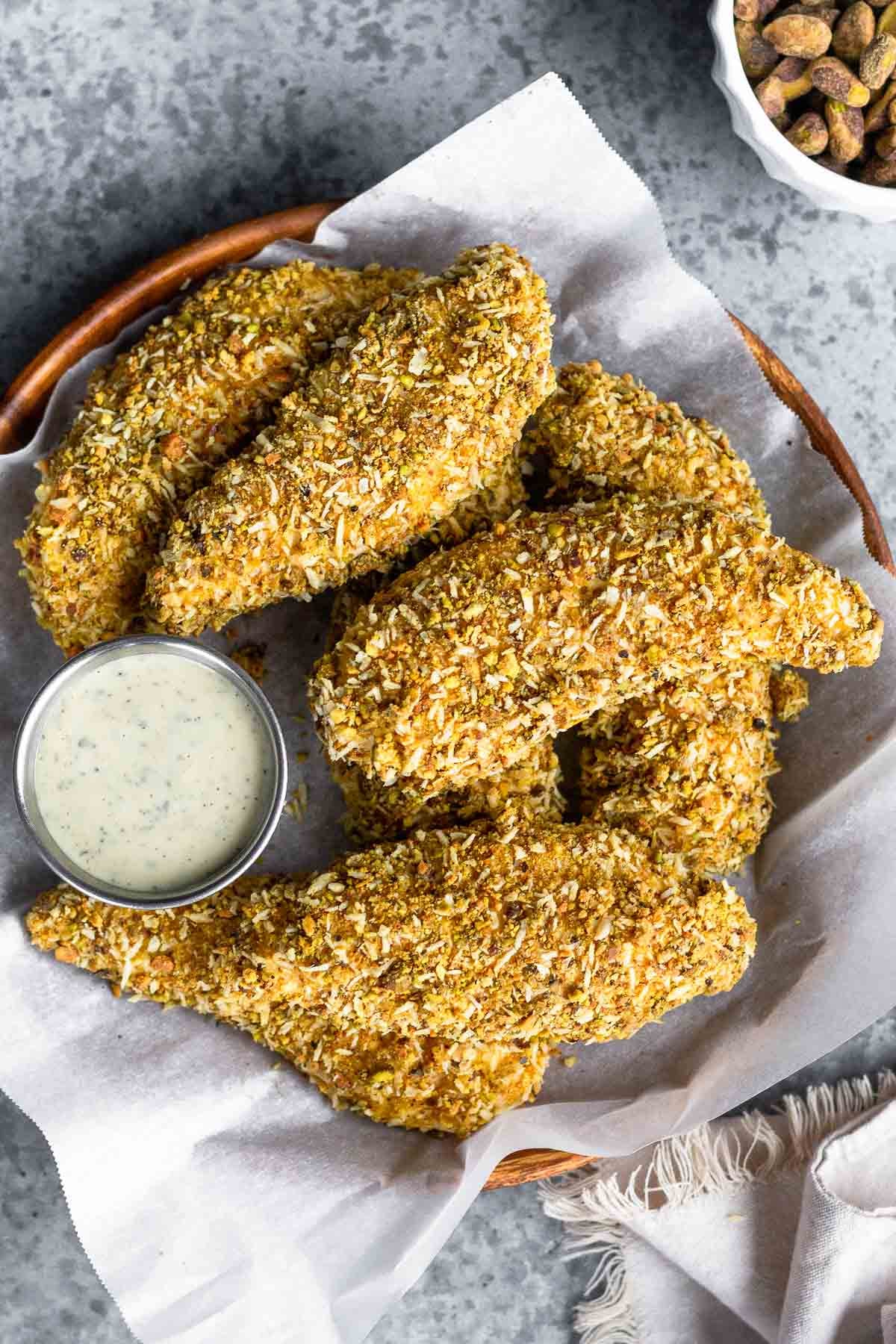 Pistachio crusted chicken tenders on a plate piled on top of each other. There is a small ramekin of ranch on the plate too. Next to the plate is a bowl of shelled pistachios and a white kitchen towel.