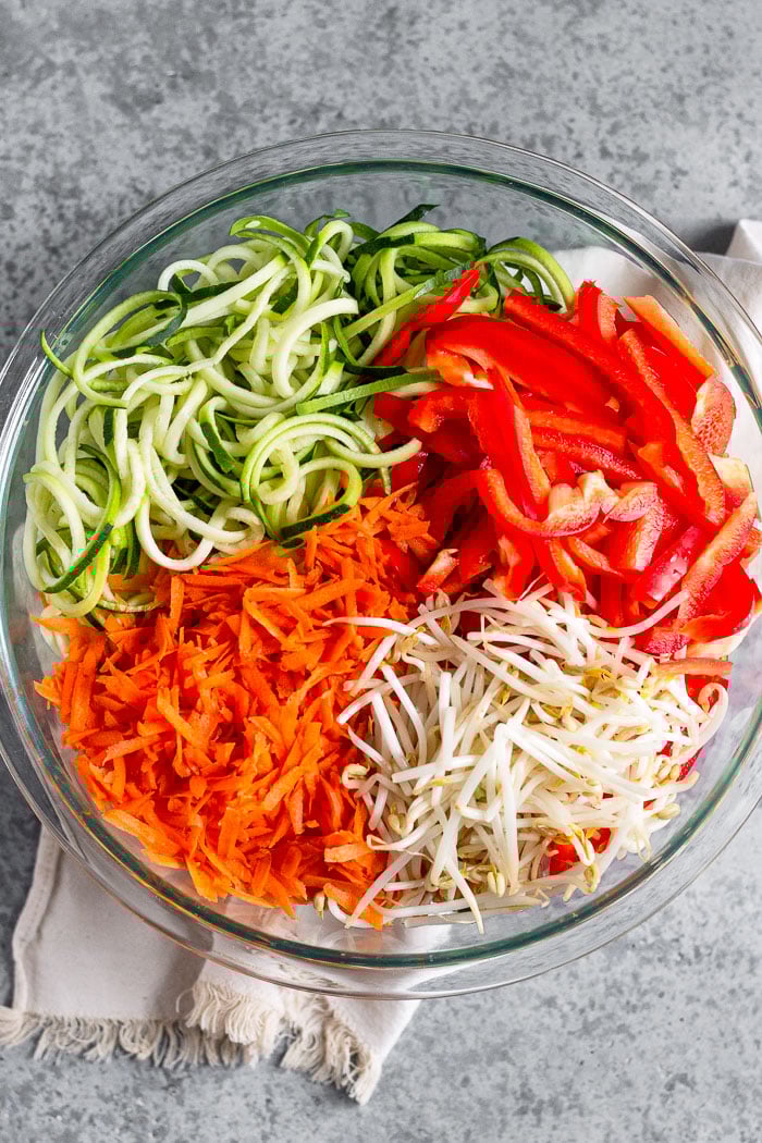 Large glass bowl of zucchini noodles, chopped bell peppers, bean sprouts, and shredded carrots.