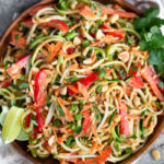 Plate filled with raw pad thai salad made from zucchini noodles, carrots, and bell peppers. It is garnished with lime wedges, cilantro, and peanuts. Next to it is a bowl of peanuts, white linen, and half a lime.