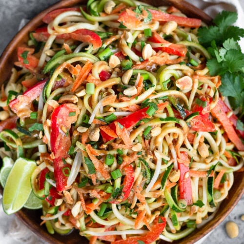 Plate filled with raw pad thai salad made from zucchini noodles, carrots, and bell peppers. It is garnished with lime wedges, cilantro, and peanuts. Next to it is a bowl of peanuts, white linen, and half a lime.