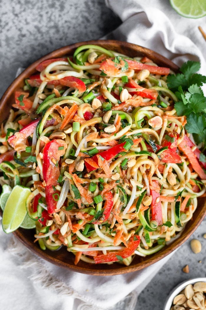 Plate filled with raw pad thai salad made from zucchini noodles, carrots, and bell peppers. It is garnished with lime wedges, cilantro, and peanuts. Next to it is a bowl of peanuts, white linen, and half a lime.