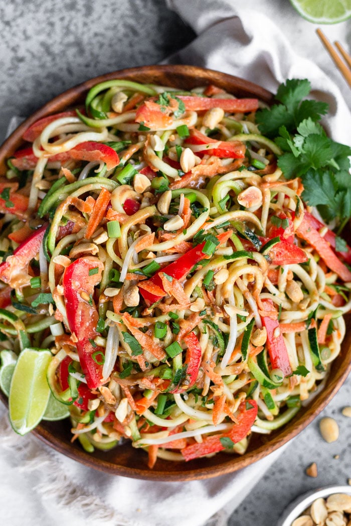 Wooden plate filled with easy vegan pad thai garnished with peanuts, lime wedges, and cilantro. It is sitting on a white linen and next to it is a bowl of peanuts, chopsticks, and half a lime.