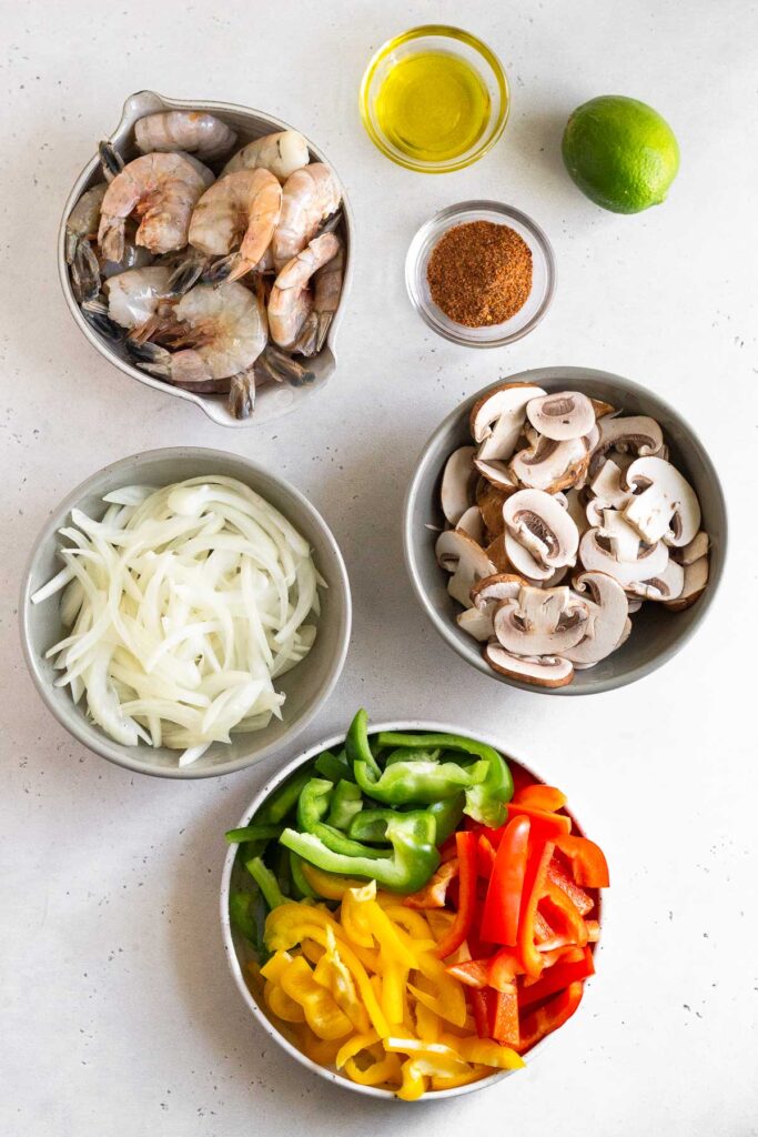 White counter of a bowl of raw shrimp, a bowl of olive oil, bowl of taco seasoning, a whole lime, a bowl of sliced mushrooms, a bowl of sliced peppers, and a bowl of sliced onions.