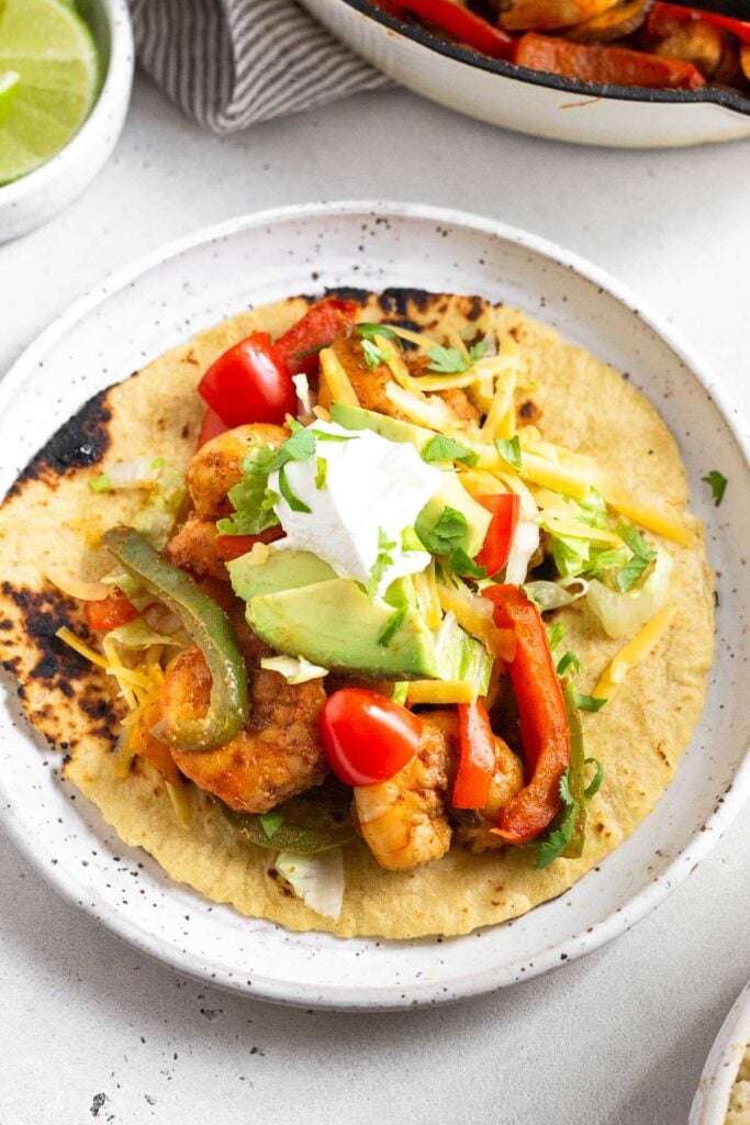 A tortilla filled with shrimp, peppers, lettuce, tomato, cheese, avocado, sour cream, and cilantro on a plate. Behind it a pan of move filling and a bowl of lime wedges.
