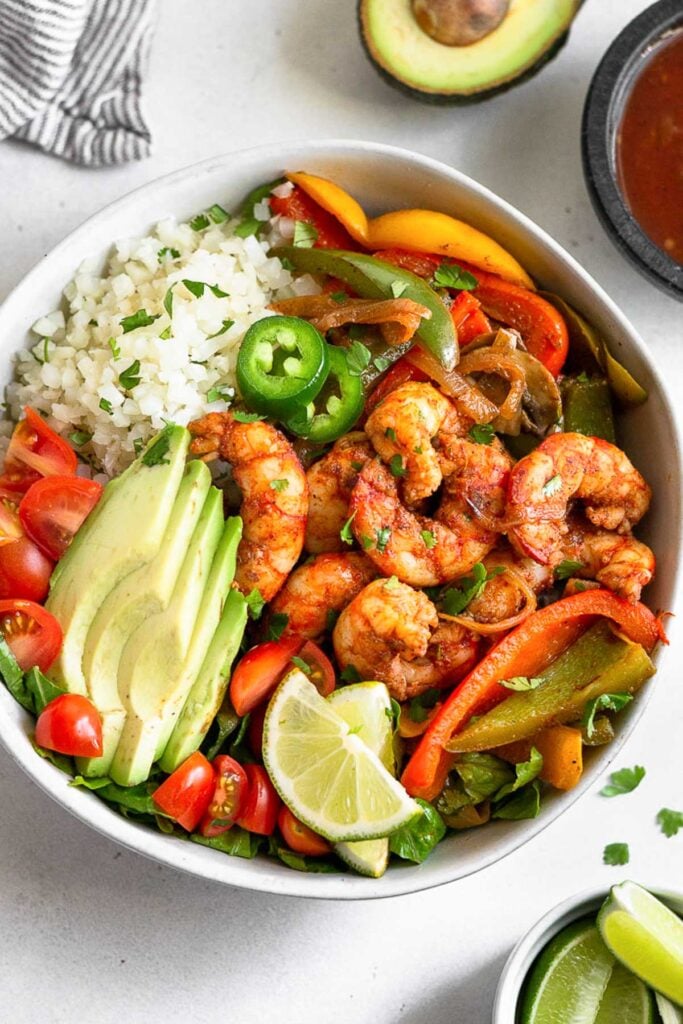 A taco bowl with cauliflower rice, sautéed peppers, onions, and mushrooms, seasoned shrimp, diced tomatoes, avocado, sliced jalapeños, and lime wedges on a bed of lettuce. Around it is half an avocado, a bowl of salsa, and a bowl of lime wedges.
