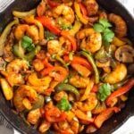 Shrimp fajitas in a large pan garnished with cilantro. The pan is sitting on a towel and around it is a bowl of rice and a bowl of limes wedges.