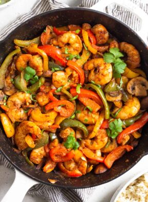 Shrimp fajitas in a large pan garnished with cilantro. The pan is sitting on a towel and around it is a bowl of rice and a bowl of limes wedges.