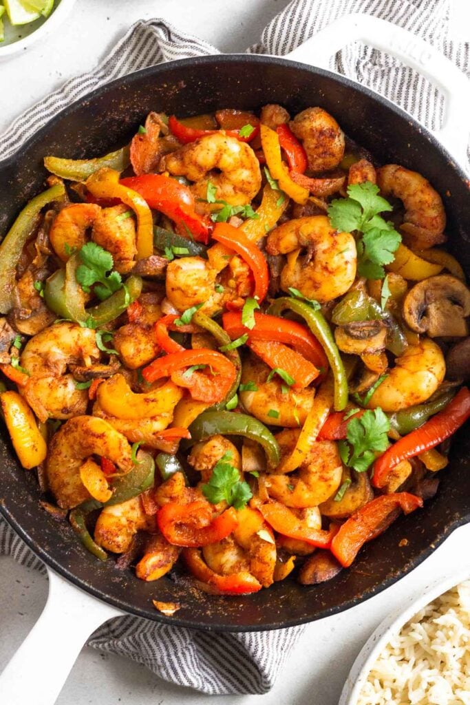 Shrimp fajitas in a large pan garnished with cilantro. The pan is sitting on a towel and around it is a bowl of rice and a bowl of limes wedges.