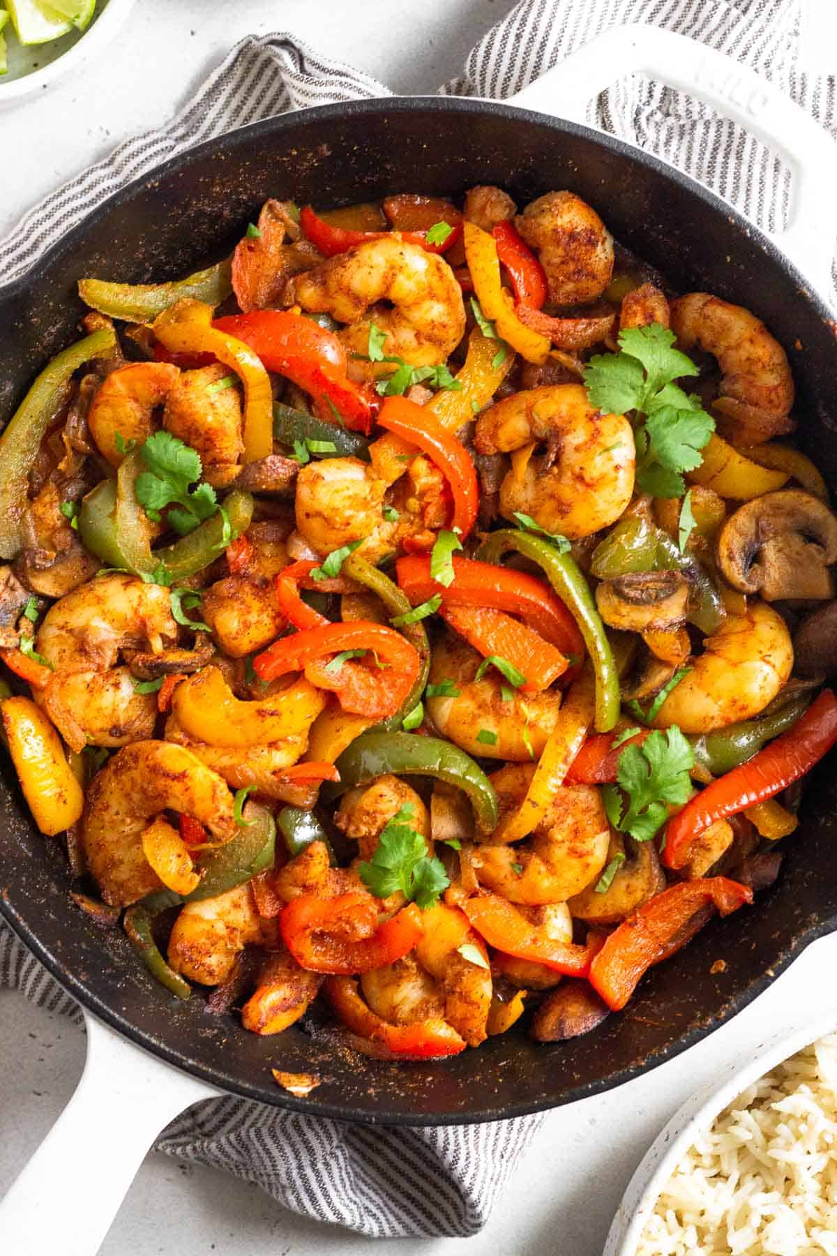 Shrimp fajitas in a large pan garnished with cilantro. The pan is sitting on a towel and around it is a bowl of rice and a bowl of limes wedges.