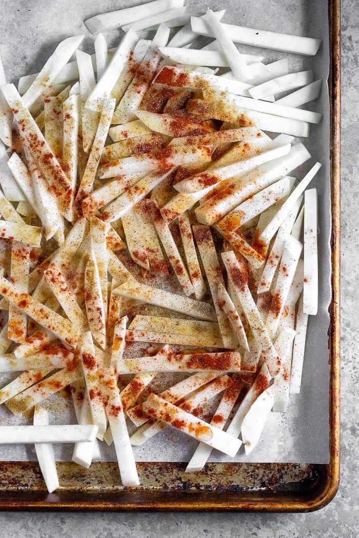 Baking sheet lined with parchment paper with turnips cut into matchsticks sprinkled with spices.