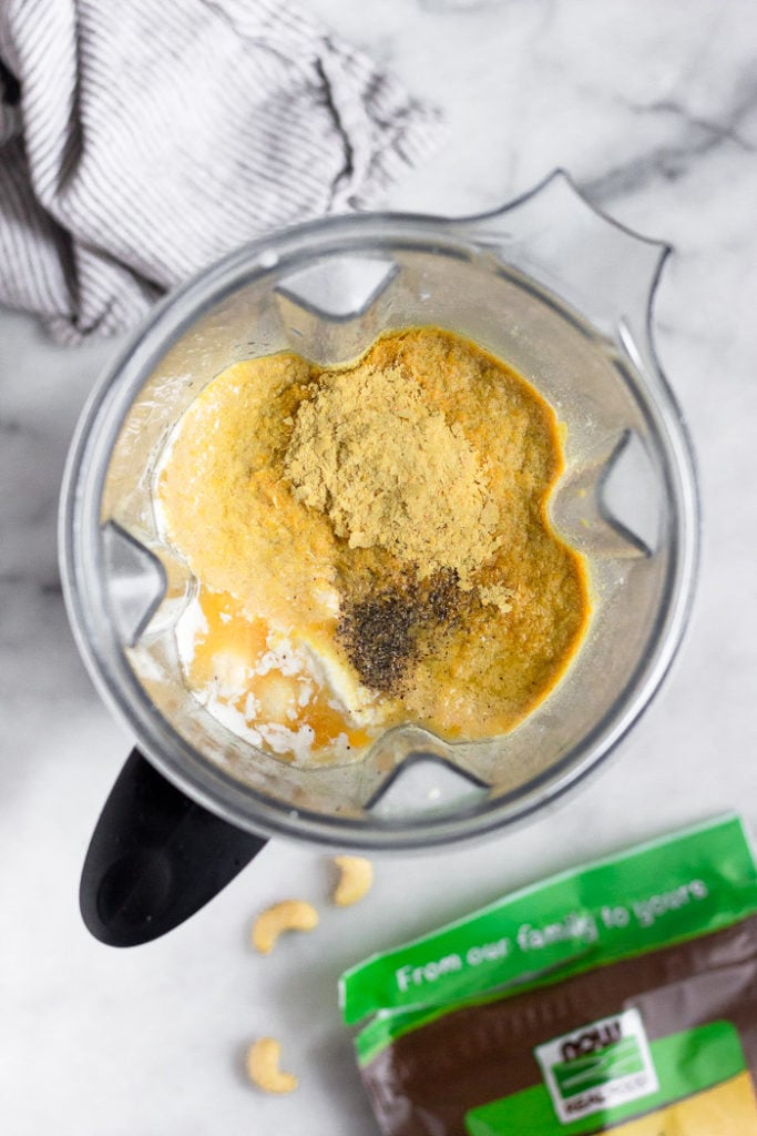 Blender filled with broth, coconut milk, cashews, nutritional yeast, and salt and pepper.