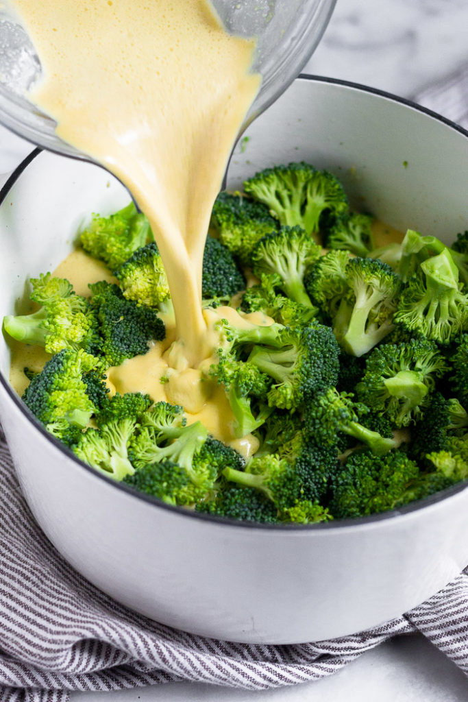 Large white dutch oven filled with fresh broccoli and a vegan cheesy broth being poured over it.