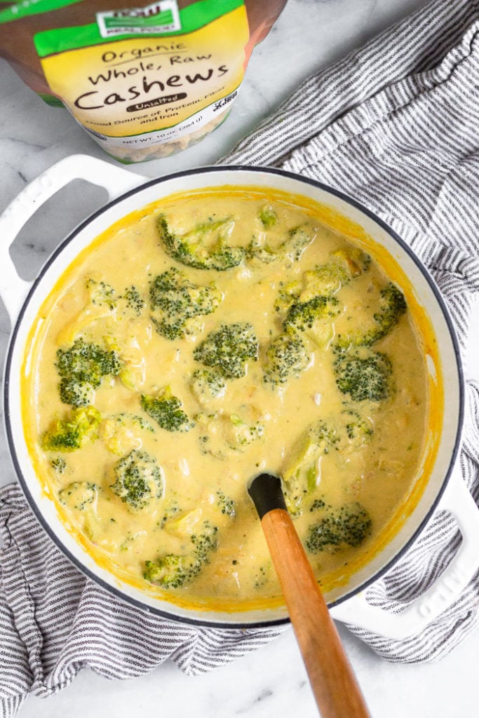 Overhead shot of easy broccoli cheese soup with a ladle coming out of it. It is sitting on a striped towel with a bag of cashews next to it.