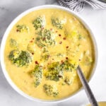 A bowl filled with vegan broccoli cheese soup topped with red pepper flakes and a spoon coming out of it. Next to it is another bowl of soup and a pot of soup.