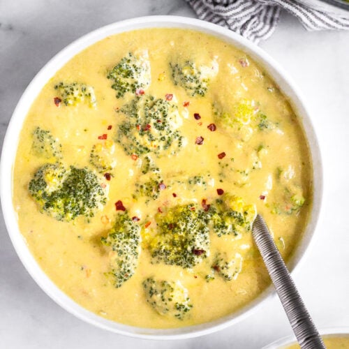 A bowl filled with vegan broccoli cheese soup topped with red pepper flakes and a spoon coming out of it. Next to it is another bowl of soup and a pot of soup.