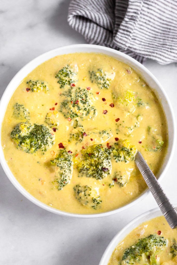 Bowl of vegan broccoli cheddar soup with a spoon coming out of it and topped with red pepper flakes. Behind it is a striped bowl and in front of it is another bowl of soup.